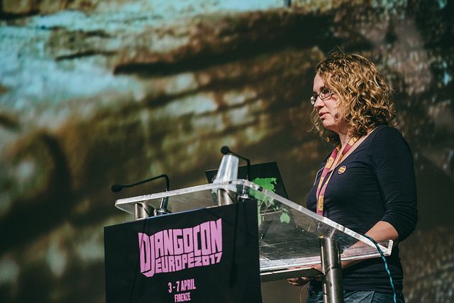 Me, woman wearing a dark top with glasses and curly blond hair, standing behind a speaker desk with the djangocon europe logo on the front.