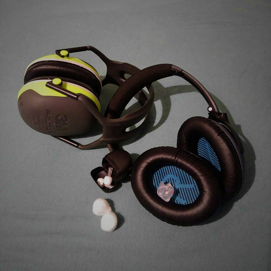 Lying on a grey surface are bose noisecanceling headphones, 3m ear defenders with a green border, custom made earplugs, earplugs with filters and wax earplugs.